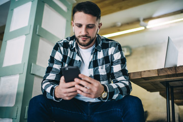 Young caucasian male in earbuds enjoying watching video on mobile phone via 4G connection and...