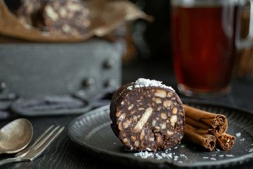 Piece of chocolate salami sprinkled with coconut and cinnamon sticks on metal plate, close-up