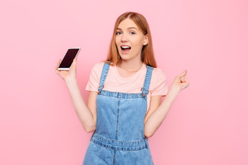 woman showing a blank smartphone screen and pointing at it while looking at the camera with her mouth open on a pink background