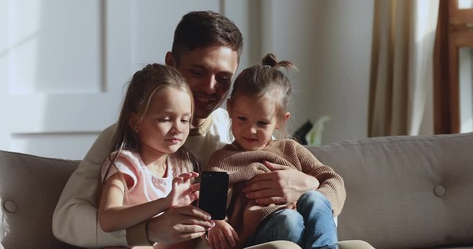 Handsome young father holding on lap little cute kids daughters, recording funny videos, taking selfie together in living room. Smiling dad showing educational mobile apps to small children at home.