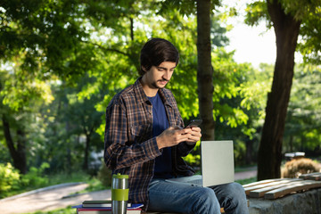Fototapeta premium Young freelancer with beard and mustache sitting with a laptop in a park and holding smartphone
