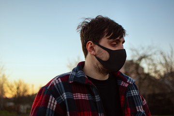 
Portrait of a guy in a black mask on a background of a sunset sky on a city street. Protective, medical mask against viruses, germs.  illness, infection. Quarantine Covid 19. Health safety concept