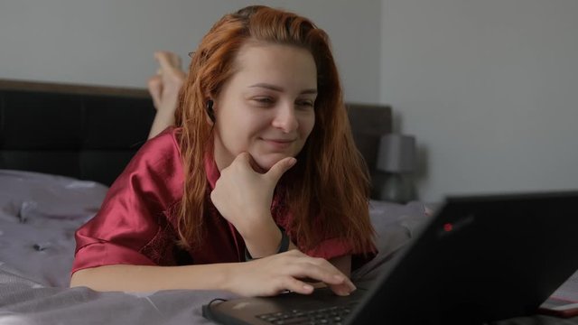 women with red and wet hair after taking a shower is lied on bed and looking for a movie while she is talking with her friend on the computer