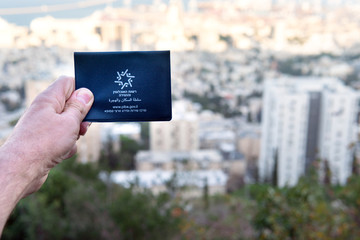 Repatriant’s hand holds a temporary Israeli passport that is issued to newly arrived returnees against the backdrop of an Israeli city Haifa