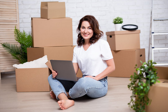 moving day concept - young woman using laptop in new house or flat surrounded with cardboard boxes