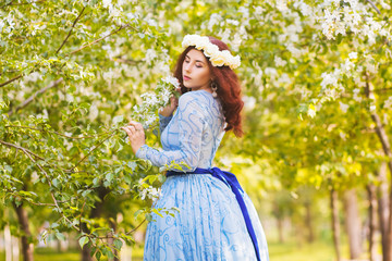 A beautiful woman in a blue puffy dress with a rim of white flowers on her head walks through the park between blooming apple trees.