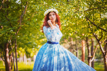 Obraz na płótnie Canvas A beautiful woman in a blue puffy dress with a rim of white flowers on her head walks through the park between blooming apple trees.