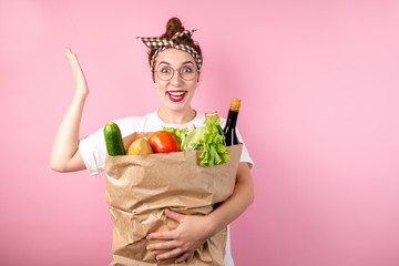 Happy housewife girl hugging a large bag of groceries on a pink background, raising her hand up in surprise