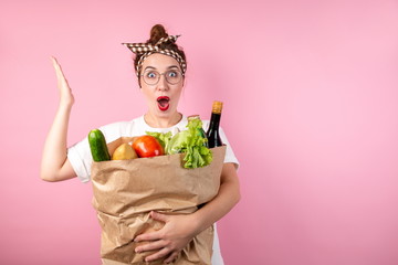 Happy housewife girl hugging a large bag of food and bottles of alcohol on a pink background raising her hand in surprise