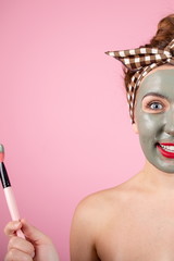 Portrait of a cute girl with a clay mask on her face holding a cosmetic brush in her hands standing on a pink background vertical photo