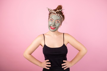 Girl in a clay mask on her face standing on a pink background with her hands on her hips looking at the camera