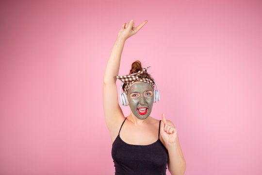 Portrait Of A Strange Girl With Red Lipstick In A Clay Mask On Her Face Listening To Music On Headphones Dancing On A Pink Background