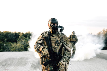 Armed soldier in a gas mask with a rifle standing on the roof of a building amid smoke and a partner. Strona protection from disorder associated with the coronavirus pandemic