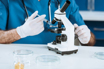 cropped view of biologist in latex gloves holding syringe near microscope