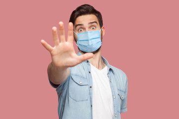 Stop, don't come any closer. . Portrait of angry young man with surgical medical mask in blue shirt standing with stop hand sign and looking at camera. indoor studio shot, isolated on pink background.