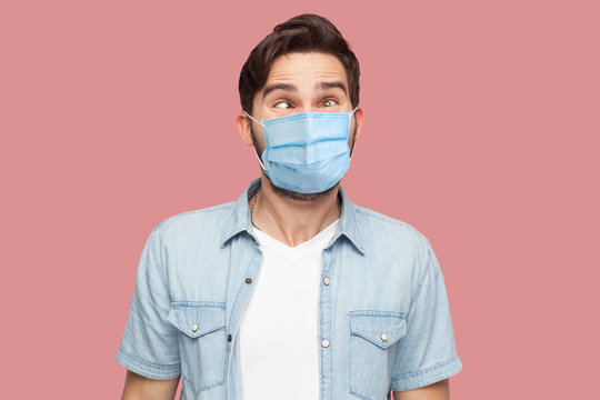 Portrait of crazy young man with surgical medical mask in blue shirt standing with crossed eyes and looking with funny comedian face. indoor studio shot, isolated on pink background.