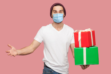 Fototapeta na wymiar Satisfied young man with surgical medical mask in white shirt and casual hat standing, holding two present box with raised arms, looking at camera. Indoor, isolated, studio shot, pink background