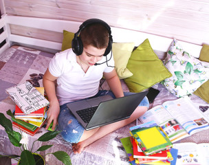 A young Caucasian, attractive boy 
in a white t-shirt and jeans shorts , in headphones is studying with a books and a laptop. Distance learning during a pandemic 2019-ncov. Insulation. 
 Education