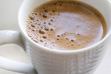 Close up of white cup of fresh coffee with foam reflecting rainbow, on light background.