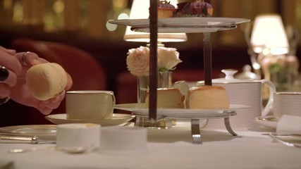 A lady's hand is picking up a piece of scone from the afternoon tea stand's lowest plate and tearing half. The camera is following her hand smoothly.