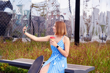 Young skater woman in blue dress making selfie by smartphone having coffee in a paper cup, sitting outdoor in a park.
