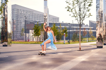 Skater woman in blue dress is training alone on longboard, doing a trick outdoor in a public park. Summer sunny day.