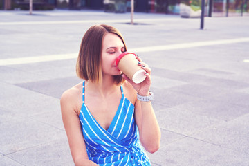 Young woman in blue dress with open shoulders having coffee in a paper cup, sitting outdoor in a park.