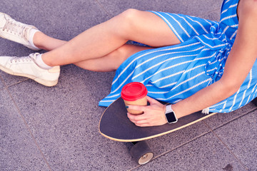 Skater woman in dress is sitting on longboard with paper cup of coffee. Close up view on hand and board.