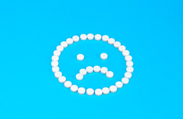 Sad face made of white round pills on a blue background. Concept of ill health or illness.