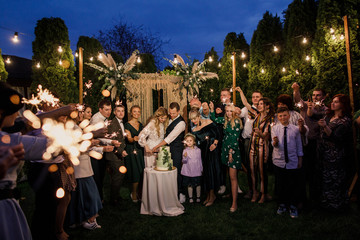 young beautiful wedding couple cut wedding cake with friends and have fun with bengal lights