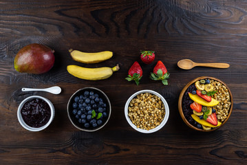 Food knolling. A wooden bowl of granola, fruits. Blueberry and banana smoothie dressing, decorated with strawberries, mangoes. Flatlay of Ingredients on dark background.