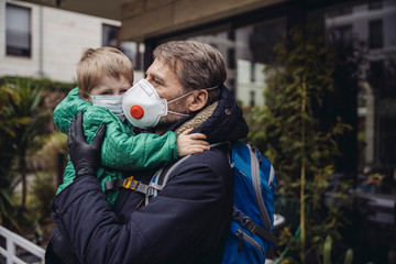 Father carrying son, both wearing protective maska, outdoors
