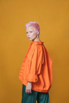 Portrait of young woman with short pink hair wearing orange jacket in front of yellow background