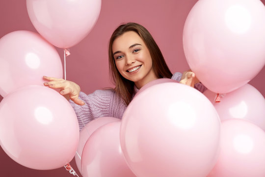 Smiling pleased girl posing with pastel pink air balloons on pink background. Beautiful happy young woman on a birthday holiday. close-up