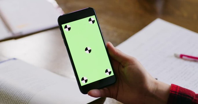 Female student looks at her green screen phone as she studies for school and takes notes as she's reading from her textbooks.