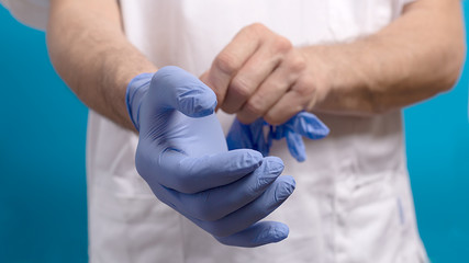 A male doctor surgeon in white uniform puts on blue gloves.