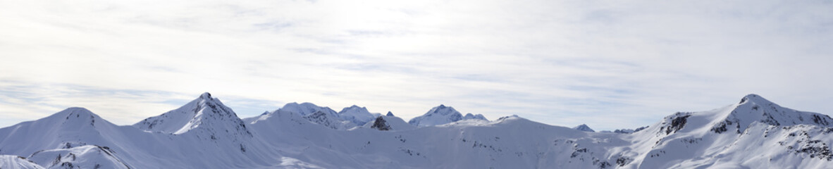 Panorama of high winter mountains with snowy slopes and sunlit cloudy sky