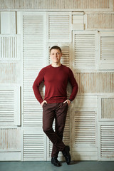Young man stands in full growth leaning against wall holds his hands in his pockets, smiling