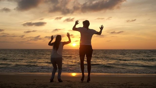 active couple silhouettes jump with joy spending time on ocean coast against waves at tropical sunset backside view slow motion