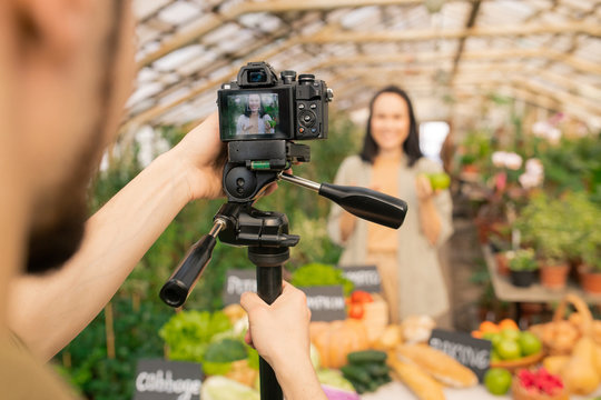 Man adjusting camera on tripod while making video about gardening with Asian farmer in greenhouse