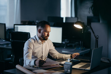 Caucasian man works alone at office in the evening and looking at laptop.