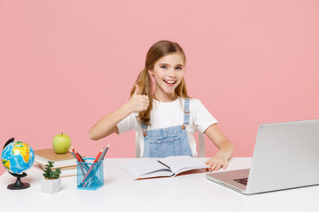 Cheerful little kid schoolgirl 12-13 years old sit study at white desk with pc laptop isolated on pastel pink background. School distance education at home during quarantine concept. Showing thumb up.