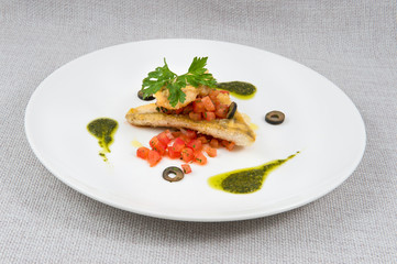 Fried fish with tomatoes, which are diced, decorated with olives, sauce with herbs, and parsley on a large round plate on a gray background