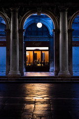 Symmetrical night shot of a closing bar in Saint Mark's square in Venice, framed by two marble columns and a round arch