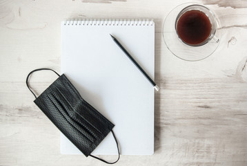 Black medical mask and notebook. Next to a cup of coffee and a pencil. Work from home. Stay at home in quarantine. Coronavirus concept