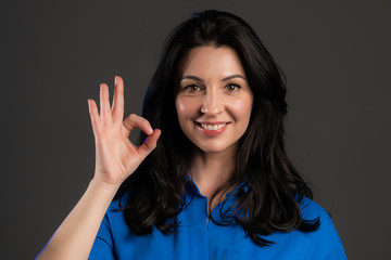 Happy adult woman on grey background. Lady with long hair in blue shirt shows Ok gesture. Winner. Success. Positive lady smiles to camera. Body language.