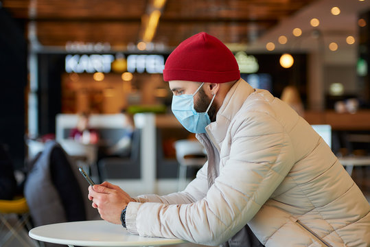 A caucasian man with a beard wearing a medical face mask to avoid the spread coronavirus (COVID-19). A guy with a surgical mask on the face using a smartphone in the mall.