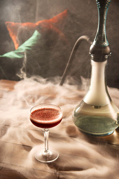 A glass of raspberry cocktail next to a hookah on a wooden table in a dark bar. Alcohol and smoking concept.