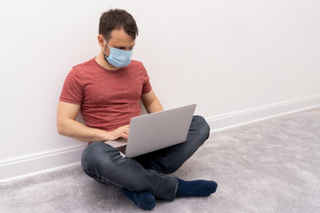 Man at self quarantine works from home in laptop at remote job, guy at protective surgical mask sits on floor. Protective measures from coronavirus.