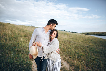 Fototapeta premium Happy couple in love hugging, kissing and smiling against the sky in field. Hat in girl's hand